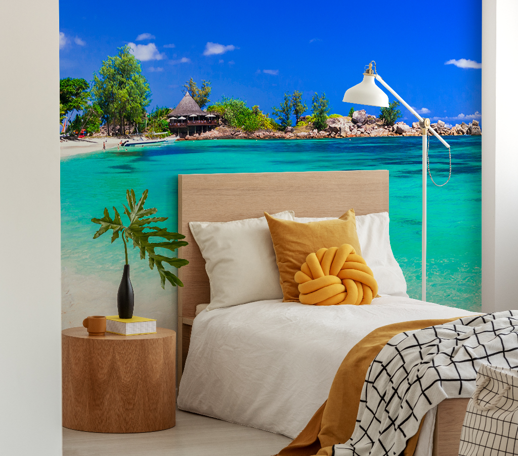 A stylish bedroom with a Decor2Go Tropical Blue Wallpaper Mural on the wall, featuring a wooden bed with beige and yellow pillows, a side table with a lamp and books, and a small plant.