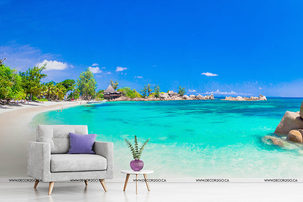 A surreal digital montage combining a Tropical Blue Wallpaper Mural from Decor2Go Wallpaper Mural with a pristine tropical beach with crystal-clear turquoise water and an interior space featuring a grey armchair and small coffee table with a plant.