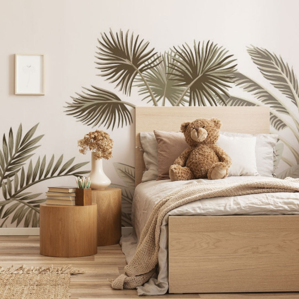 A cozy bedroom with a single bed featuring a teddy bear, a bedside table with books and a vase of flowers, against a Decor2Go Wallpaper Mural leaf-patterned wall. A minimalist art frame hangs above.