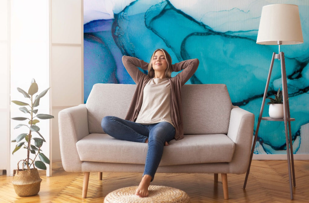 Living Room - A woman relaxes on a sofa with her hands behind her head in a cozy living room, surrounded by plants and vibrant Blue Ocean Waves Wallpaper Mural from Decor2Go.