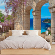 Bedroom - A cozy bedroom with a wooden bed frame and white bedding exudes rustic elegance, complemented by a Tuscany Veranda View Wallpaper Mural from Decor2Go. This large wall mural showcases a stone archway reminiscent of a Tuscany veranda, offering an overlook of the vibrant blue sea and lush green island, framed by blooming purple wisteria.