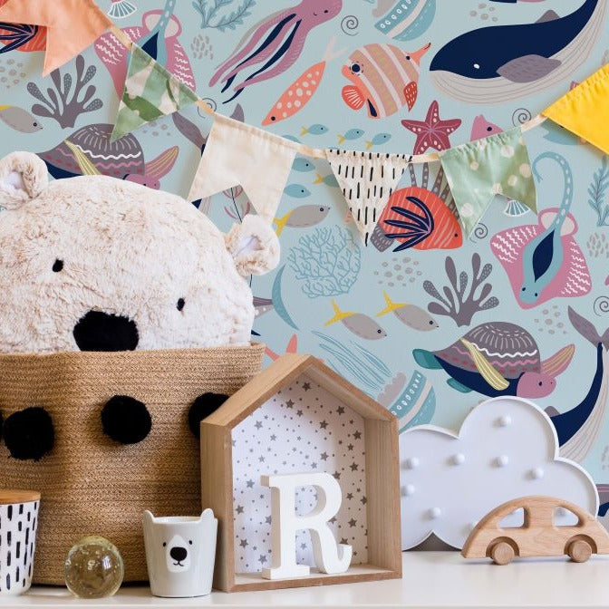 A cheerful children's room corner with a whimsical Decor2Go Wallpaper Mural featuring colorful fish and whales. A plush teddy bear, wooden toys, and decorative items are neatly arranged.