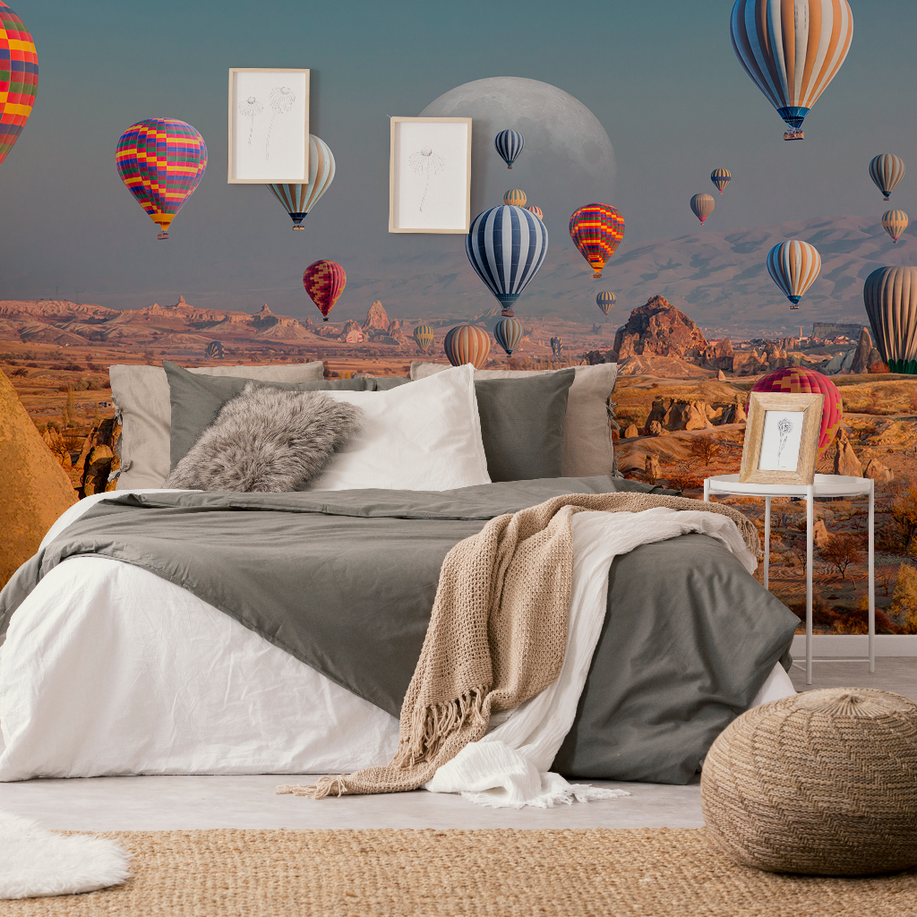 Bedroom - A cozy bedroom features a large bed with gray and white bedding, a tan knit throw, and a fluffy pillow. The wall showcases Decor2Go's Skyward Dreams Wallpaper Mural, depicting vibrant hot air balloons floating over rock formations under a moon.