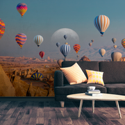 Living Room - A living room with a dark sofa, colorful cushions, and a round wooden table with a cup. The wall features Decor2Go's Skyward Dreams Wallpaper Mural, showcasing vibrant hot air balloons soaring over majestic rock formations beneath a large moon in the desert sky.