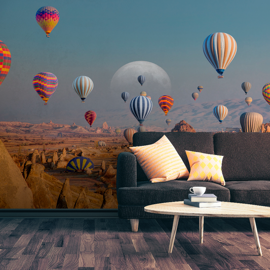A living room with a dark sofa, colorful cushions, and a round wooden table with a cup. The wall features Decor2Go's Skyward Dreams Wallpaper Mural, showcasing vibrant hot air balloons soaring over majestic rock formations beneath a large moon in the desert sky.