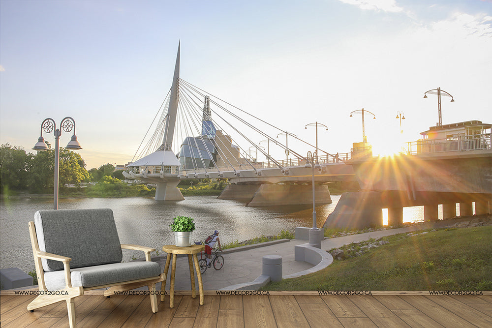 Living Room - An outdoor living room setup by a river at sunset, featuring a modern sofa and chair, overlooking the **Urban Bridge Wallpaper Mural** from **Decor2Go Wallpaper Mural**, with a small bicycle and potted plant nearby.