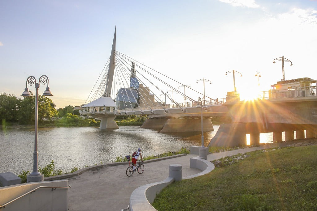 A person riding a bicycle near a river at sunset, with the Decor2Go Wallpaper Mural Urban Bridge Wallpaper Mural and a city skyline in the background. Sunlight casts a warm glow across the scene.