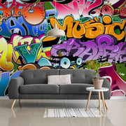 Living Room - A vibrant living room with a wall covered in colorful Urban Colors Wallpaper Mural featuring various shapes and the word "music". A modern grey sofa with a pillow, a small white side table, and a potted