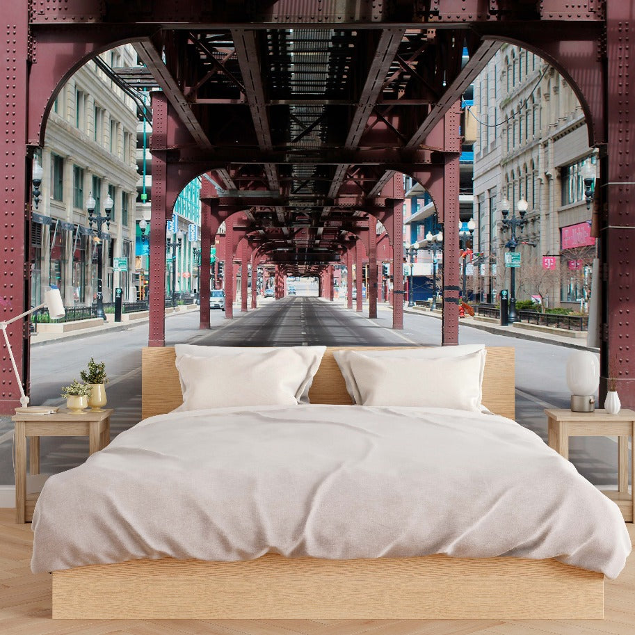 A surreal image of a neatly made bed with a beige comforter and white pillows, placed on a city street under a dark metallic Decor2Go Wallpaper Mural bridge structure, with buildings lining the empty street.