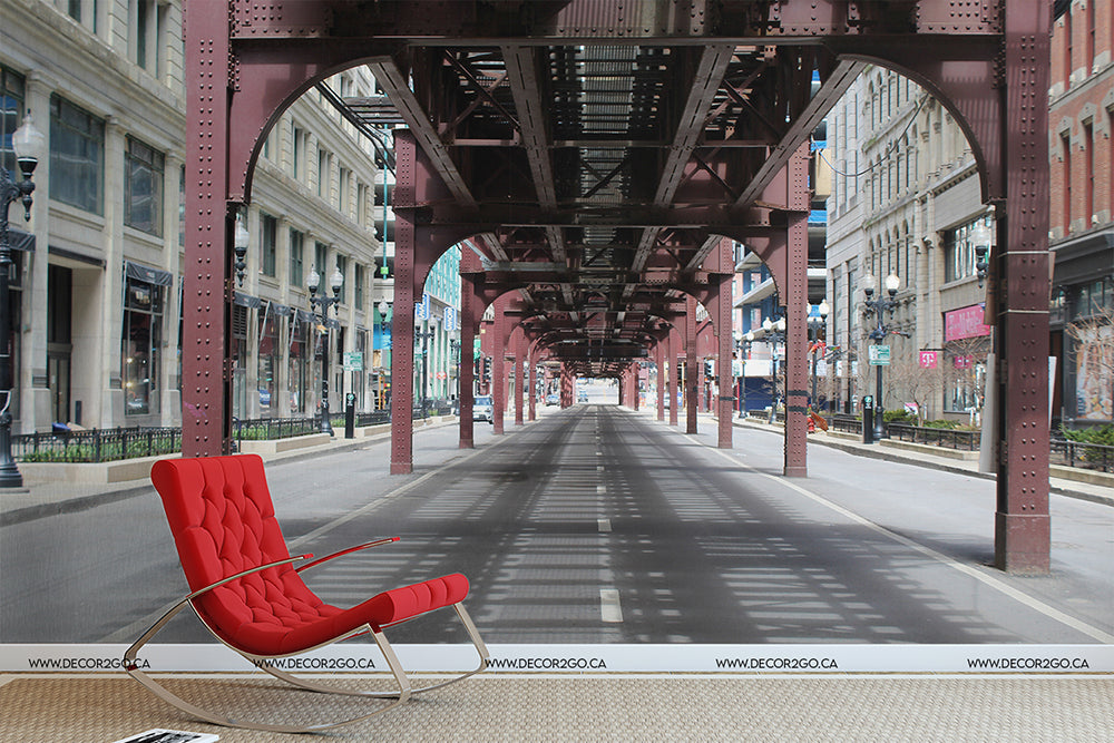 A red modern chair positioned in the center of an empty street in Queens NYC, under an urban railway track. Light casts long shadows on the concrete, enhanced by a Decor2Go Wallpaper Mural nearby, giving a stark