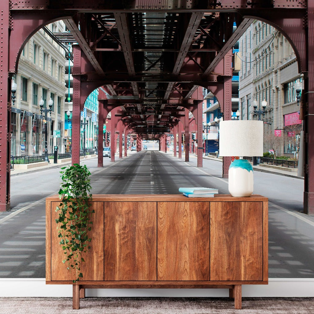 A Decor2Go Wallpaper Mural with a plant and a lamp on top sits incongruously on an empty city street under a tall urban railway bridge structure, contrasting urban architecture with home decor.