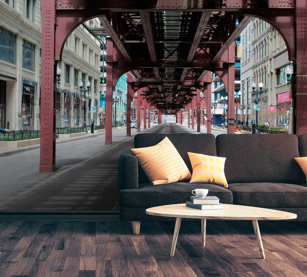A surreal living room setup under an elevated train track in Queens NYC street view includes a dark gray sofa, colorful cushions, a white coffee table with a cup, and a hardwood floor featuring the Urban Railway Wallpaper Mural by Decor2Go Wallpaper Mural.