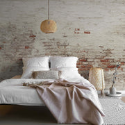 Bedroom - A cozy bedroom featuring an unmade bed with white bedding and a beige throw, against a rustic exposed brick wall enhanced by Decor2Go Wallpaper Mural. A wicker pendant light, a tall lantern, and simple