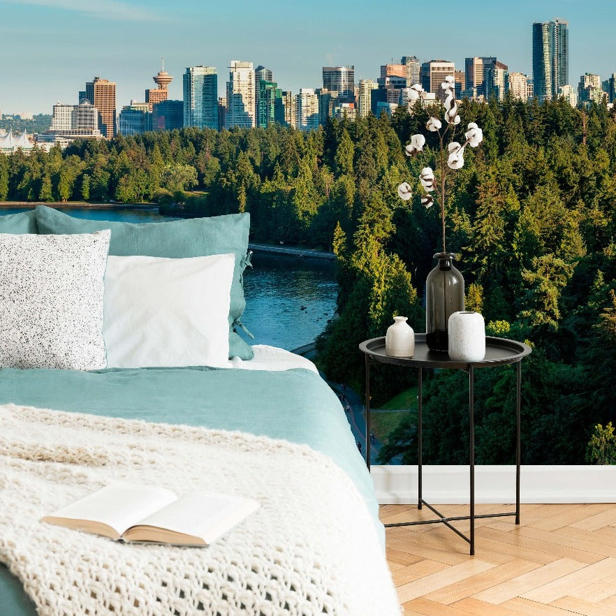 A cozy outdoor bedroom set-up with a neatly made bed and plush pillows on a balcony overlooking a scenic view of a lush park and the Decor2Go Wallpaper Mural Vancouver Summer Skyline Wallpaper Mural under a clear blue sky.