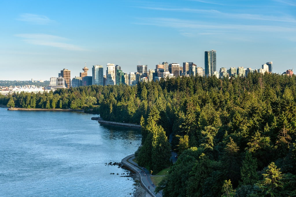 A picturesque view of the Decor2Go Wallpaper Mural Vancouver Summer Skyline Wallpaper Mural behind a lush green forest with a serene river in the foreground, under a clear blue sky.