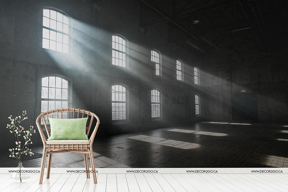 An empty industrial loft with large, architectural windows casting beams of natural sunlight across the floor, featuring a single rattan chair with a green cushion on the right foreground, and a Decor2Go Wallpaper Mural in Versailles design hanging on the back wall.
