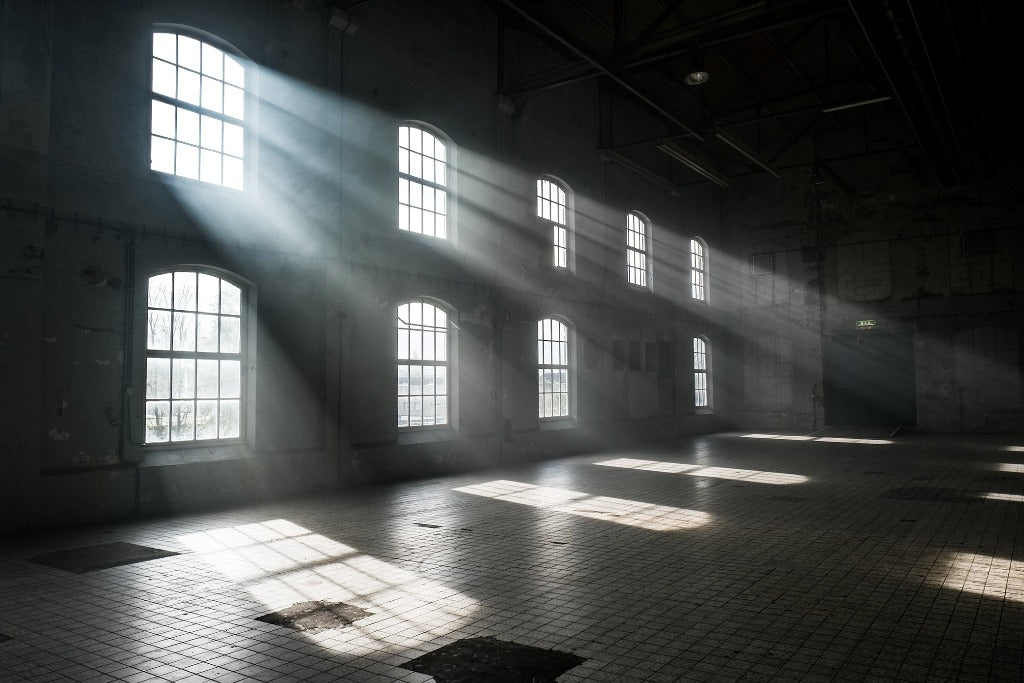 Sunlight streams through tall, architectural windows in an empty, dusty room highlighting beams of light across the shadowy interior. (Decor2Go Wallpaper Mural)