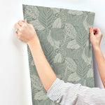 A person is hanging a sheet of York Wallcoverings Vinca Eucalyptus Wallpaper Green (60 Sq.Ft.) with a green and cream leaf pattern on a white wall. The individual, wearing a striped long-sleeve shirt and a ring on their left hand, effortlessly manages the durable non-woven material that is easy to hang and remove.