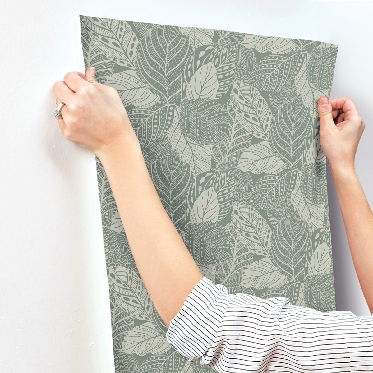 A person with a striped shirt is hanging Vinca Glint Wallpaper Metallic (60 Sq.Ft.) by York Wallcoverings with a leaf pattern on a wall. The wallpaper, made of durable non-woven material, features various leaves in shades of green and beige. The person is holding the wallpaper by its top corners.