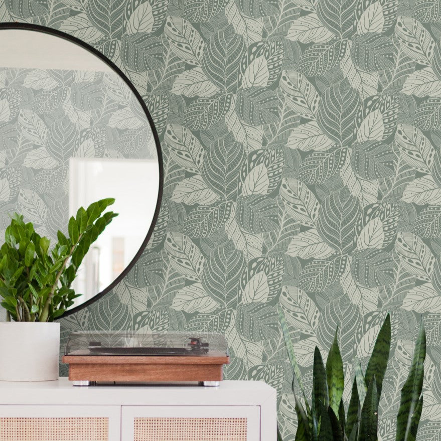 A stylish room corner featuring a Vinca Eucalyptus Wallpaper Green (60 Sq.Ft.) by York Wallcoverings, a circular mirror, a record player on a white cabinet, and plants including a potted green plant and a taller plant with dark green leaves.