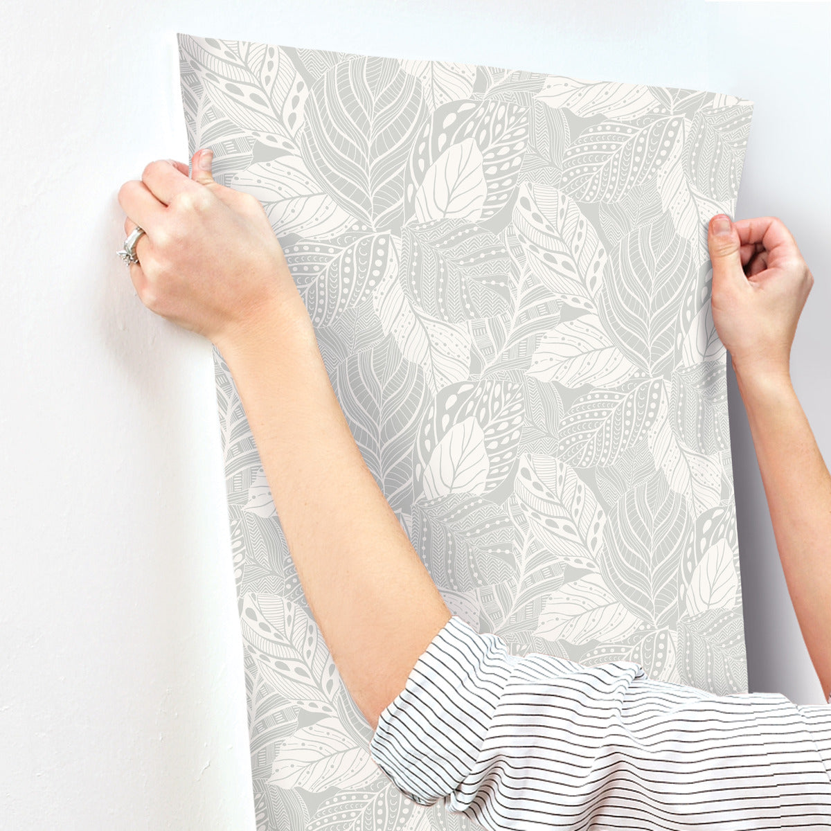 Person, wearing a striped long-sleeve shirt, affixes a sheet of Vinca Smokey Blue Wallpaper Blue (60 Sq.Ft.) by York Wallcoverings to a wall. The individual's hands are positioned at the top corners, smoothing the easy to hang and remove wallpaper into place.