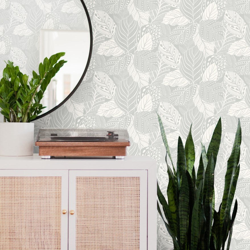 A modern living room corner featuring a white cabinet with coral-colored mesh doors, topped with a turntable and a potted plant. A round mirror hangs on the wall with York Wallcoverings Vinca Eucalyptus Wallpaper Green (60 Sq.Ft.).