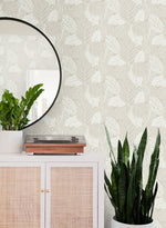 A decorative setting with a white sideboard featuring rattan doors and a round mirror above. On the sideboard, there's a potted plant and a record player. Another tall plant stands beside it, against light-colored York Wallcoverings Vinca Smokey Blue Wallpaper Blue (60 Sq.Ft.) that's easy to hang and remove.