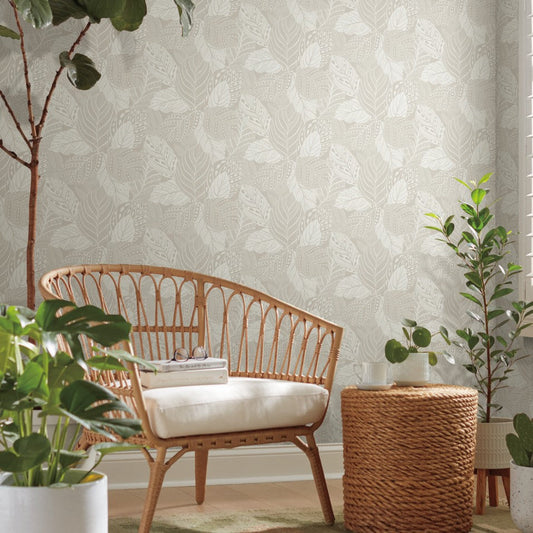 A serene room featuring a rattan chair with a white cushion, a wicker side table, and various potted plants. The walls showcase Vinca Glint Wallpaper Metallic (60 Sq.Ft.) by York Wallcoverings in a light-colored leaf pattern. Sunlight filters in through a window, casting soft light over the cozy setting.