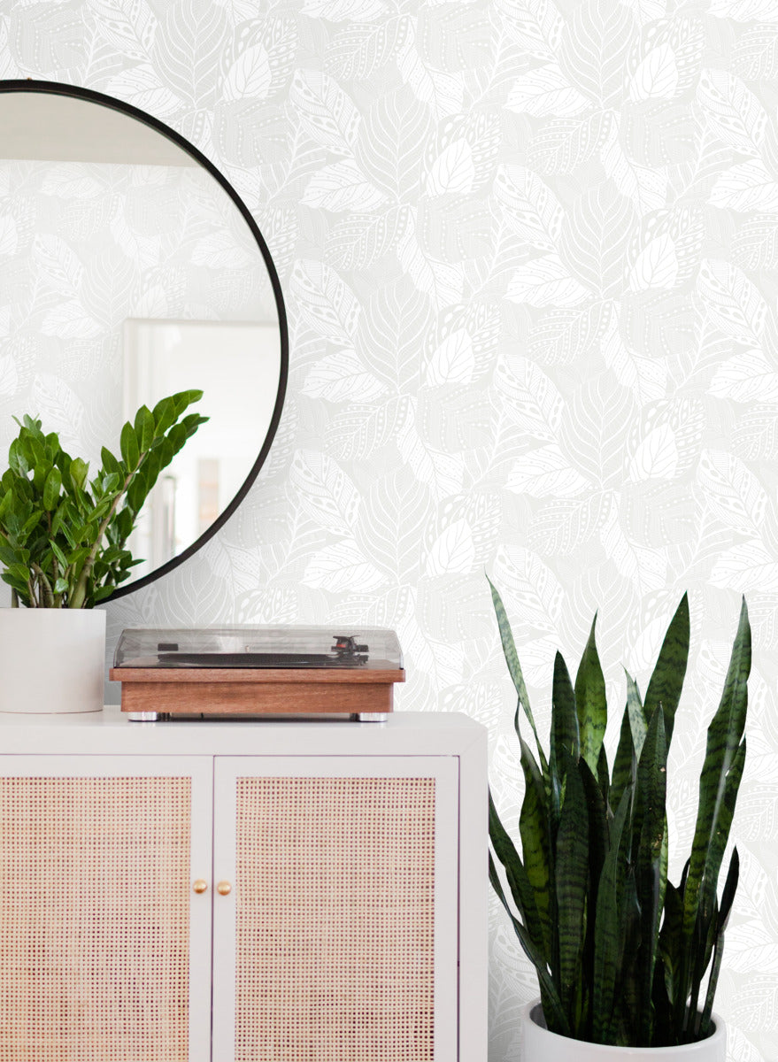 A modern interior scene featuring a white cabinet with woven front panels, topped with a potted plant and a wooden object. Above, a round wall mirror reflects part of the room. Next to the cabinet is a tall potted snake plant. The backdrop is York Wallcoverings Vinca Glint Wallpaper Metallic (60 Sq.Ft.) with its textural canopy of vines.