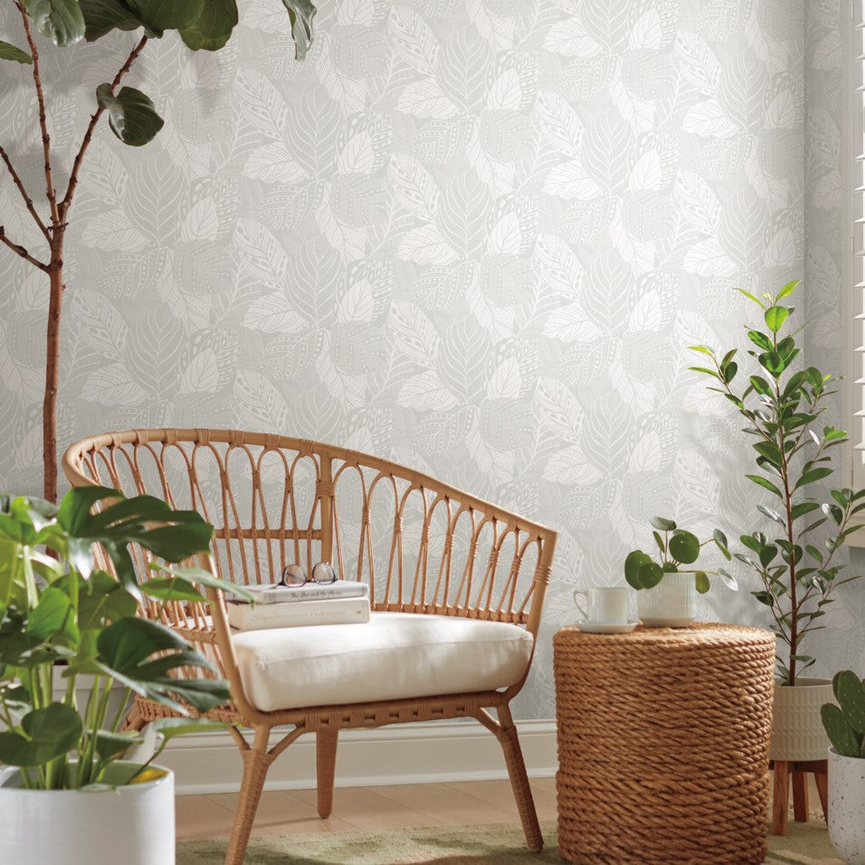 An airy room featuring a rattan bench with a light cushion, beside a woven basket with a potted plant. The room has large Vinca Eucalyptus Wallpaper Green (60 Sq.Ft.) from York Wallcoverings and additional green plants.
