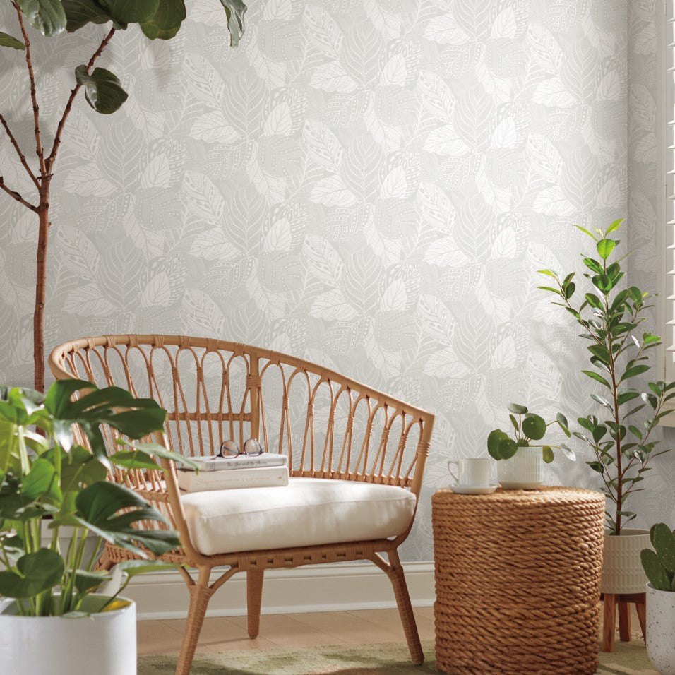 A cozy sitting area with a rattan loveseat featuring white cushions, a woven round coffee table holding a white teapot and cup, and a stack of books. The background boasts York Wallcoverings' Vinca Smokey Blue Wallpaper Blue (60 Sq.Ft.), with a large potted plant on the left and smaller greenery on the right.