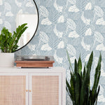 A stylish room corner with a white cabinet featuring a coral pink fabric panel, a wooden turntable on top, and a round mirror. There's a large potted plant next to the cabinet against easy to hang and remove York Wallcoverings Vinca Eucalyptus Wallpaper Green (60 Sq.Ft.).