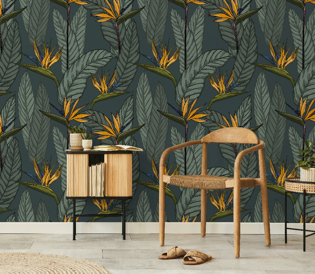 A room featuring Decor2Go Wallpaper Mural Vintage Botanical Garden Wallpaper Mural with bird of paradise flowers and large leaves. There's a wooden chair with a woven seat, a small side table holding books, and a pair of slippers on the floor. This classic decorative piece creates an overall tropical theme.