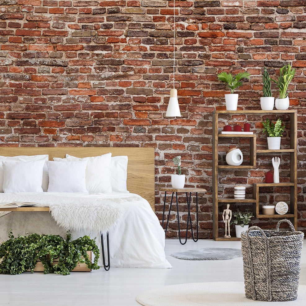 A cozy, rustic bedroom with Decor2Go Wallpaper Mural's Washed Classic Brick Wall Wallpaper Mural, featuring a bed with white linens, a wooden headboard, and a book on the bed. A shelving unit displays plants and decor.