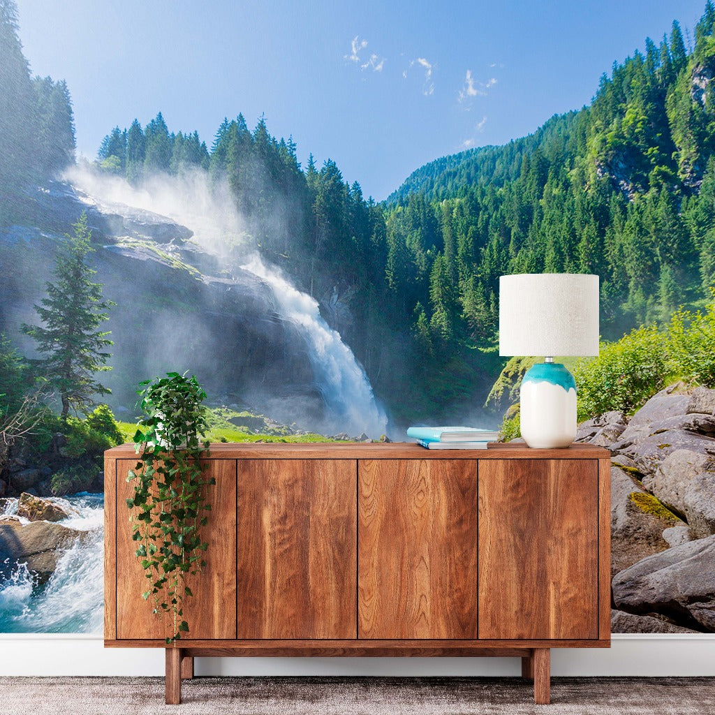 A wooden sideboard with a lamp and books in a room featuring a Water Spring or Natural Falls wallpaper mural of natural falls, trees, and a river in a lush green forest setting by Decor2Go Wallpaper Mural.