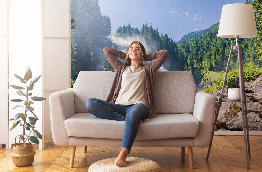 A woman relaxes on a sofa with her eyes closed and hands behind her head, in a bright living room featuring a large window with a Decor2Go Wallpaper Mural of Water Spring or Natural Falls.