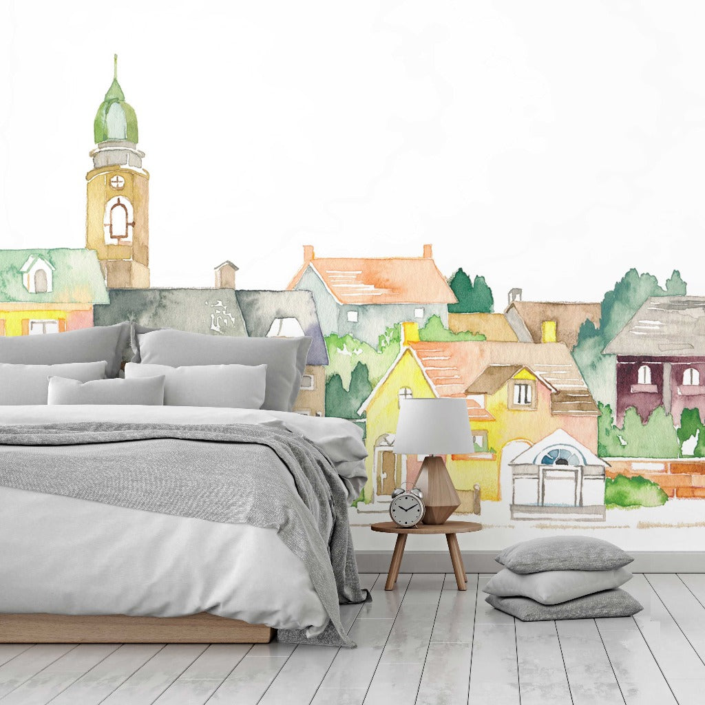 A cozy child's room with white bedding and wooden side table, in front of a Watercolor Castle Wallpaper Mural depicting a colorful, watercolor-style village with quaint houses and a tower from Decor2Go Wallpaper Mural.