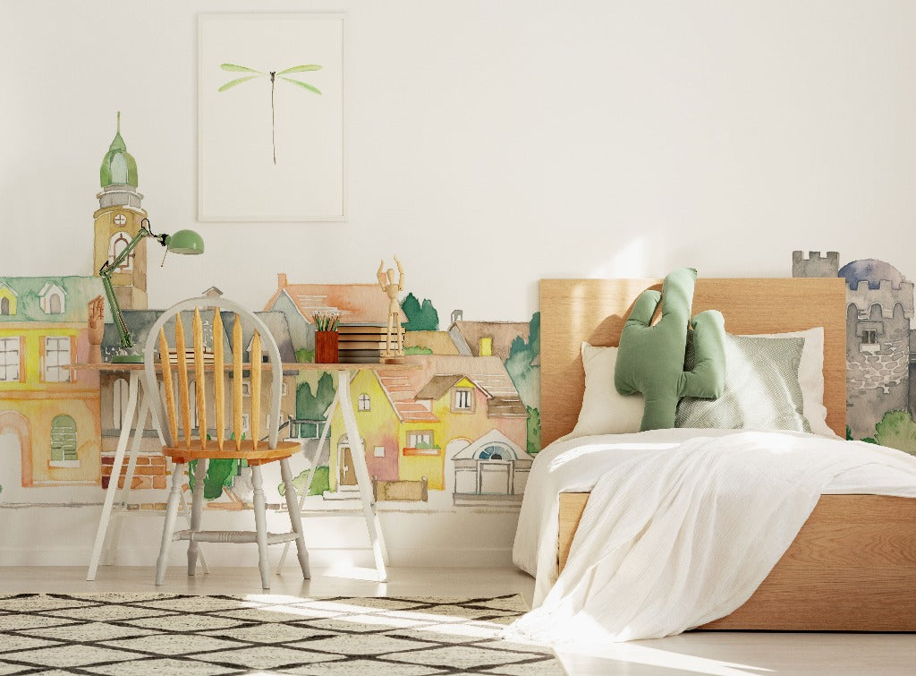 A brightly lit children's bedroom with a whimsical Decor2Go Wallpaper Mural of a watercolor castle on the wall, a wooden bed with green pillows, a chair, desk with lamp, and framed dragonfly art.