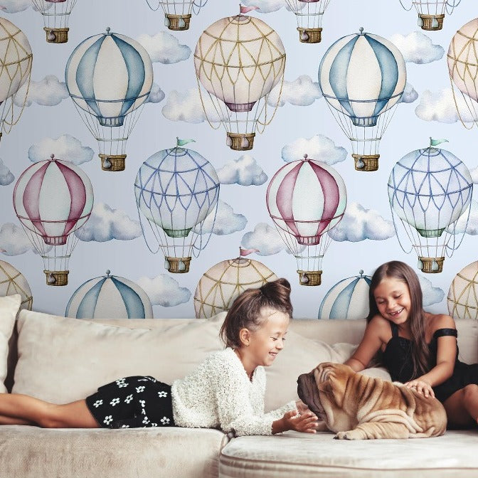 Two young girls and a large dog play joyfully on a couch in a room with walls decorated with whimsical Watercolor Hot-Air Balloons Wallpaper Mural from Decor2Go Wallpaper Mural.