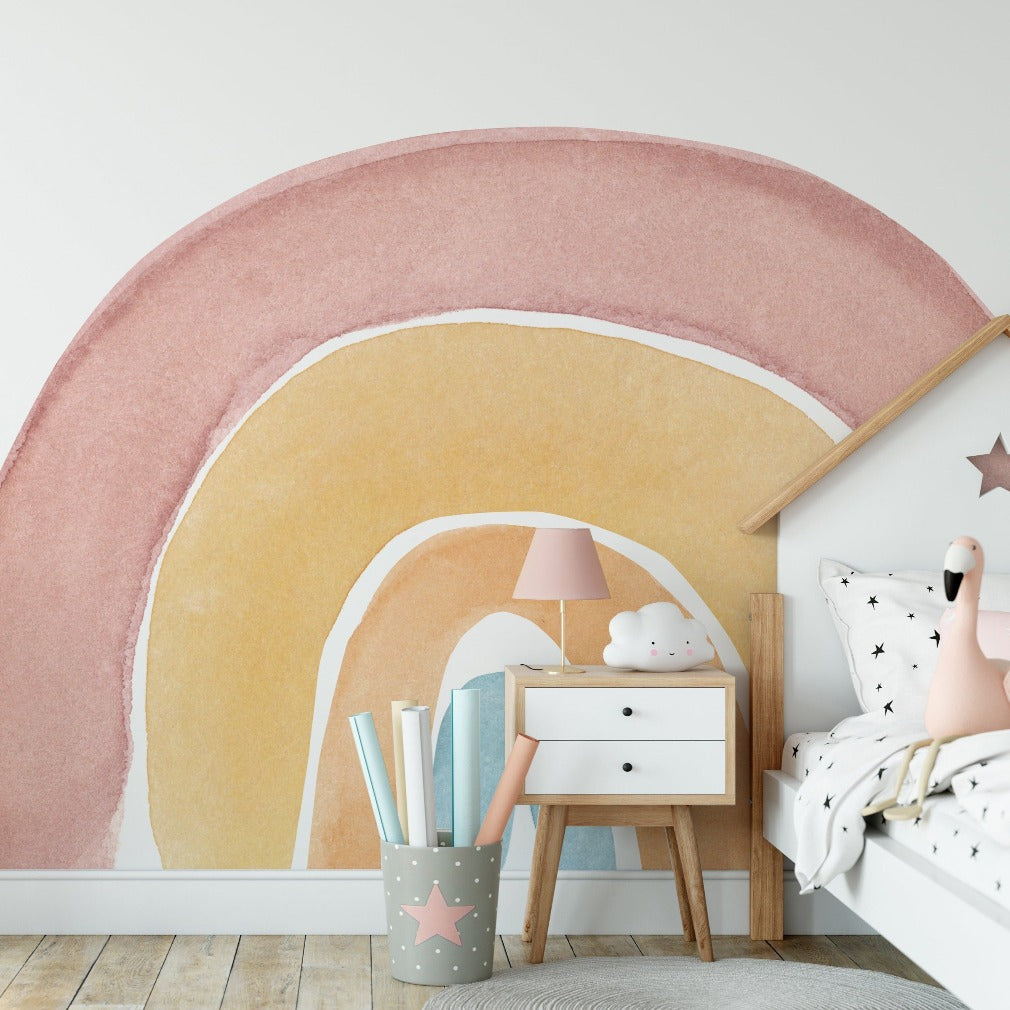 A modern children's bedroom with a large Watercolor rainbow Wallpaper Mural from Decor2Go Wallpaper Mural on the wall. Features include a wooden bed with star-patterned bedding, a white side table, books, a lamp, and a toy.
