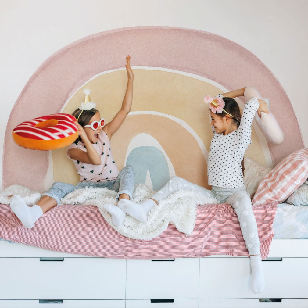 Kids Room - Two young girls having a pillow fight on a bed designed like an eye, adorned with Decor2Go Wallpaper Mural's Watercolor rainbow Wallpaper Mural, evoking a cheerful atmosphere in the children's room.
