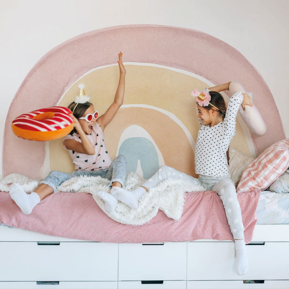 Two young girls having a pillow fight on a bed designed like an eye, adorned with Decor2Go Wallpaper Mural's Watercolor rainbow Wallpaper Mural, evoking a cheerful atmosphere in the children's room.