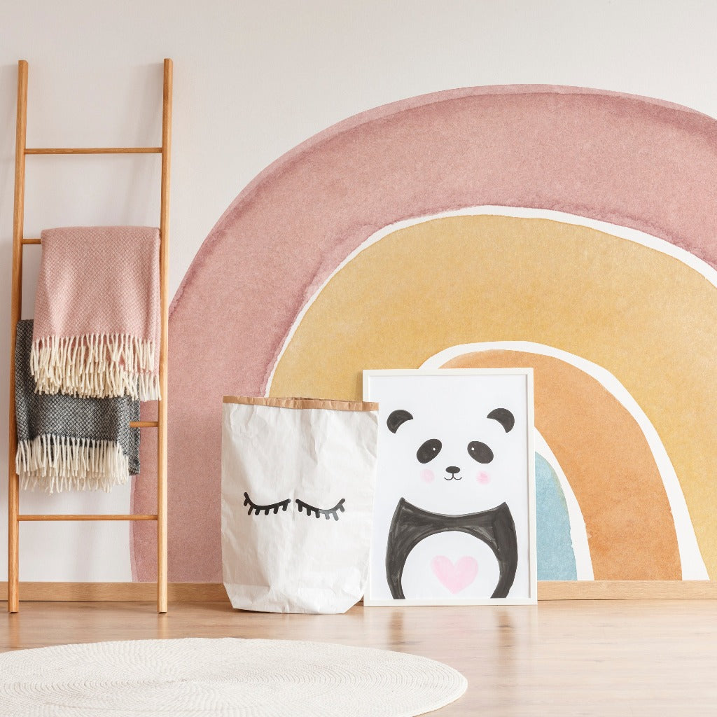 A cozy room corner featuring a wooden ladder with draped blankets, a pastel Watercolor Rainbow Wallpaper Mural from Decor2Go Wallpaper Mural, storage bags with face designs, and a framed panda painting on a white rug.