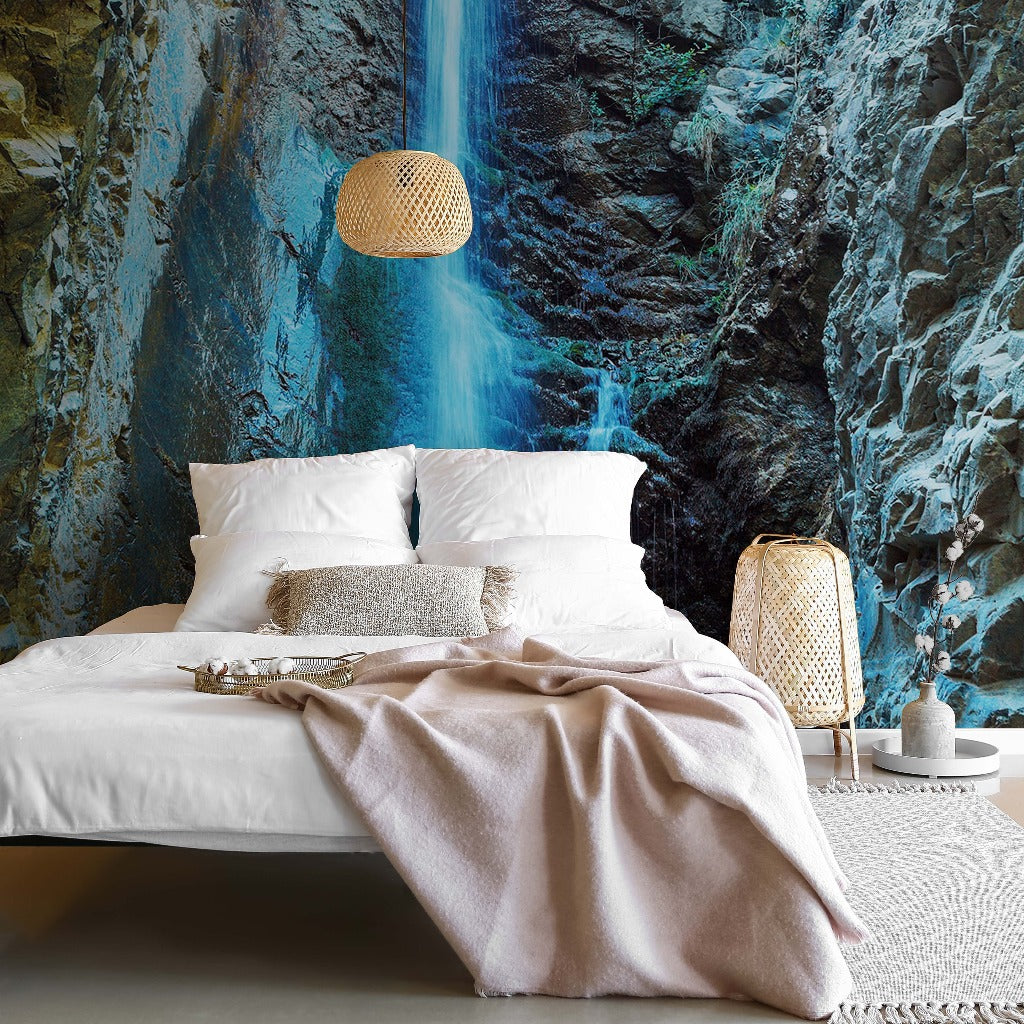 A modern bedroom featuring a large bed with white linen and a beige throw, against a striking Decor2Go Wallpaper Mural of rocky cliffs and an underground lake. Wicker lamps add to the natural aesthetic.