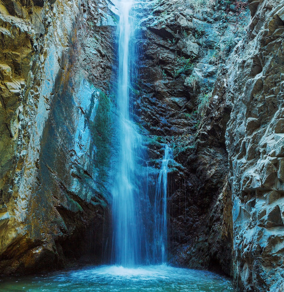A serene "Waterfall Cave Wallpaper Mural" from Decor2Go Wallpaper Mural cascades down a rocky cliff into a tranquil blue pool, surrounded by rugged canyon walls adorned with patches of green foliage and vibrant indigo touches.