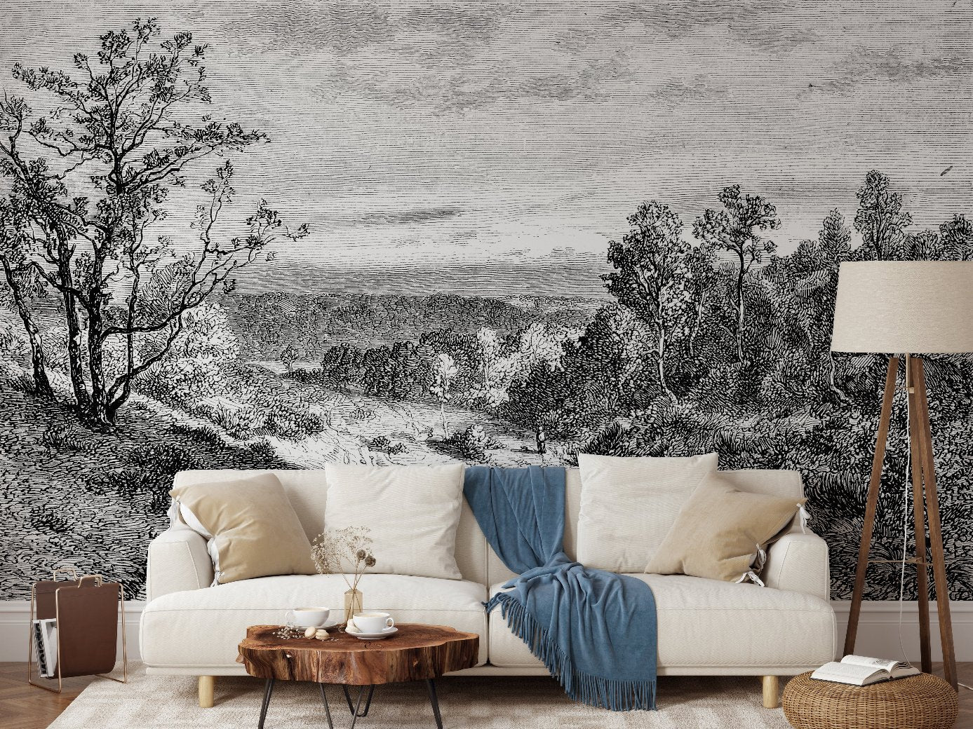 A living room with a sofa, blue throw, and white cushions. In front stands a wooden stump coffee table, complemented by a floor lamp on the right. The wall is adorned with Decor2Go Wallpaper Mural's Whispering Trees Wallpaper Mural showcasing trees and hills in black and white. A woven basket rests on the left.