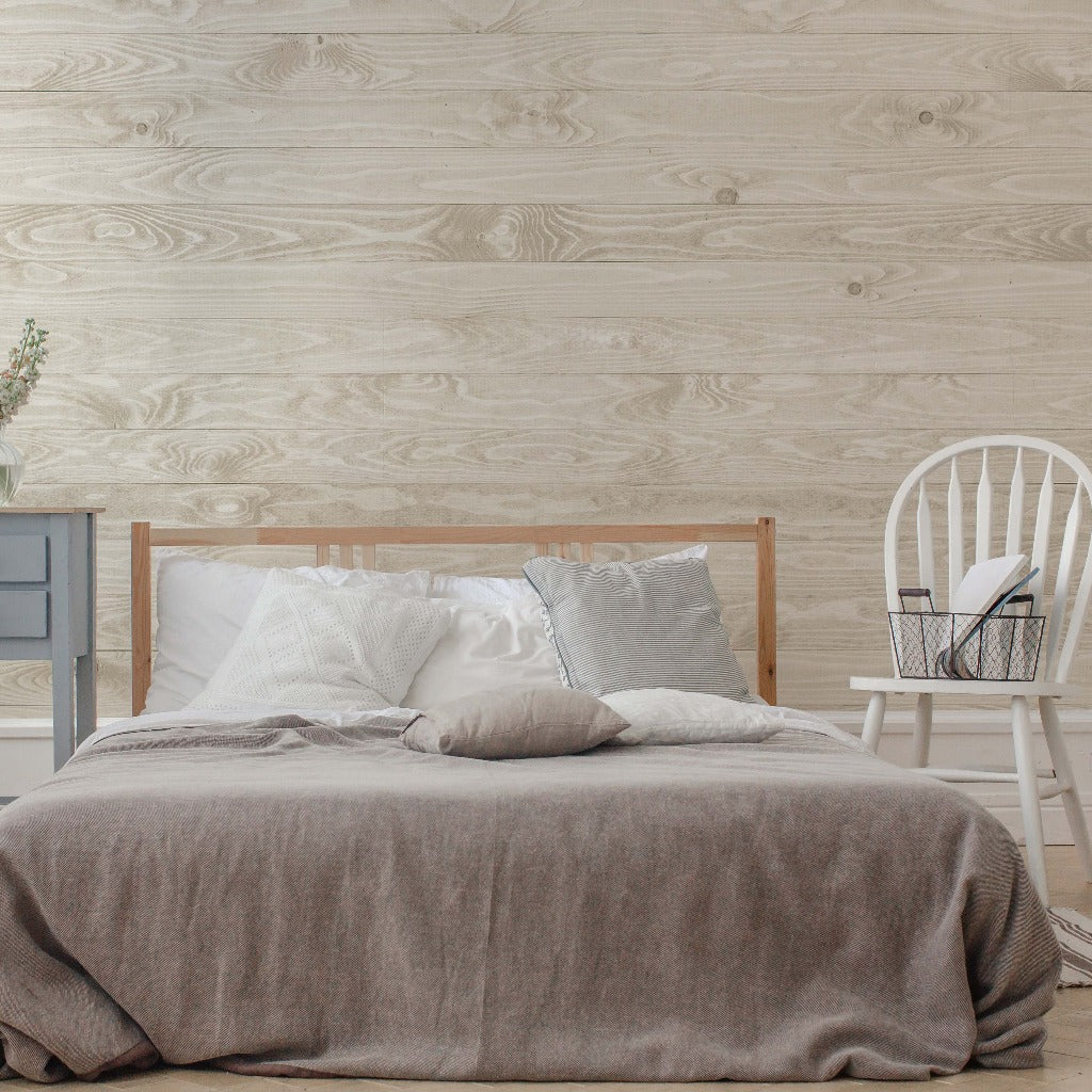 A cozy bedroom featuring a neatly made bed with gray bedding, a wooden headboard, a gray nightstand, and a white chair with a book against a Decor2Go Wallpaper Mural.