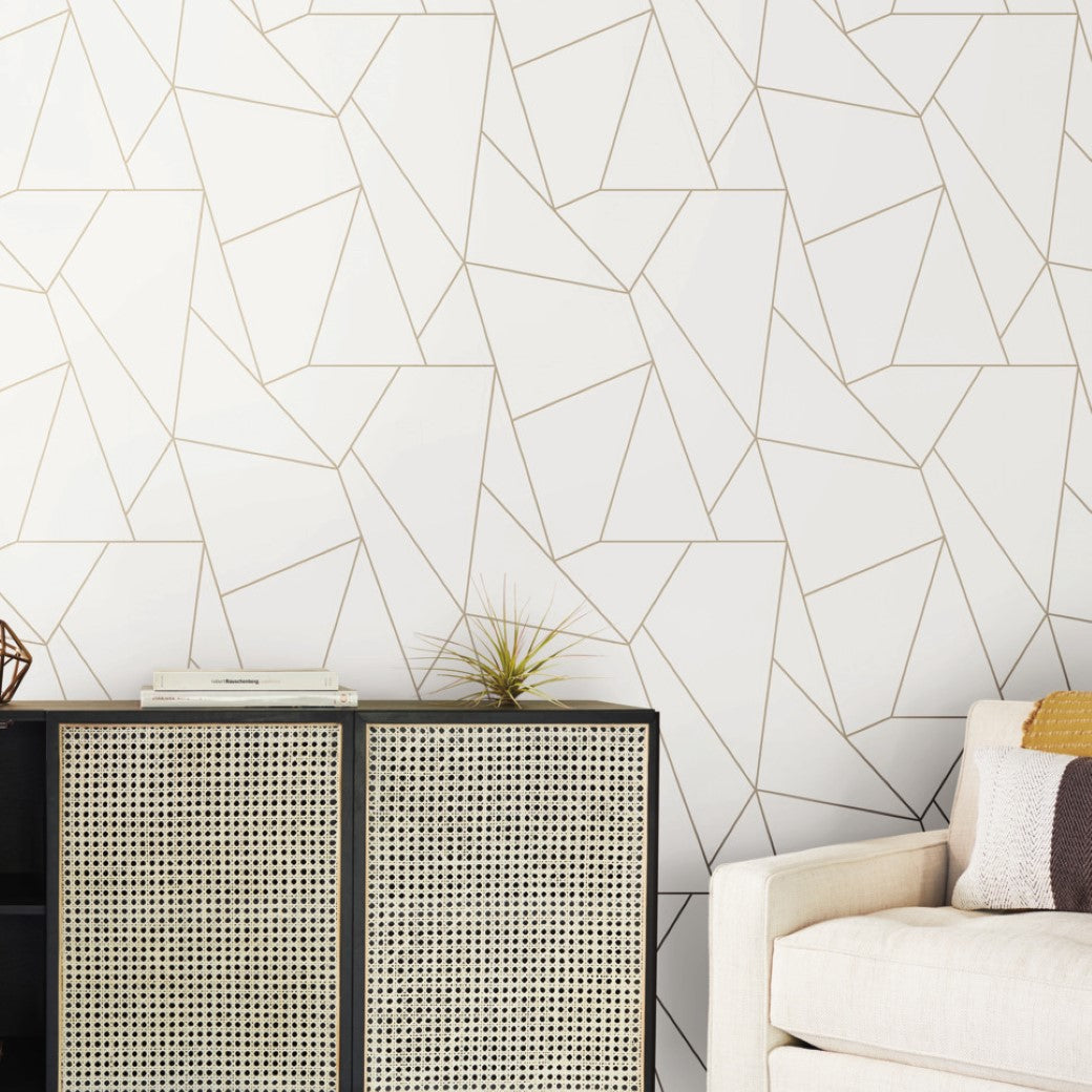 A modern living room features York Wallcoverings' White & Gold Nazca Wallpaper, displaying a geometric pattern. A black cabinet with woven rattan doors from the Modern Metals Collection holds books and a plant. On the right, a beige couch with patterned pillows is partially visible.