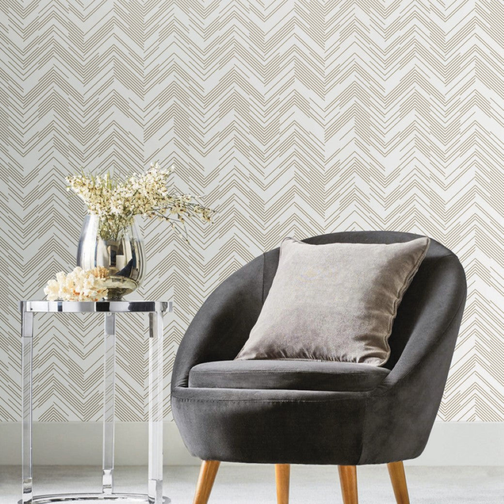 A modern living room exudes elegance with a gray padded chair, cushion, and a silver round side table holding a metallic vase of white flowers, all beautifully complemented by York Wallcoverings' White & Gold Polished Chevron Wallpaper.