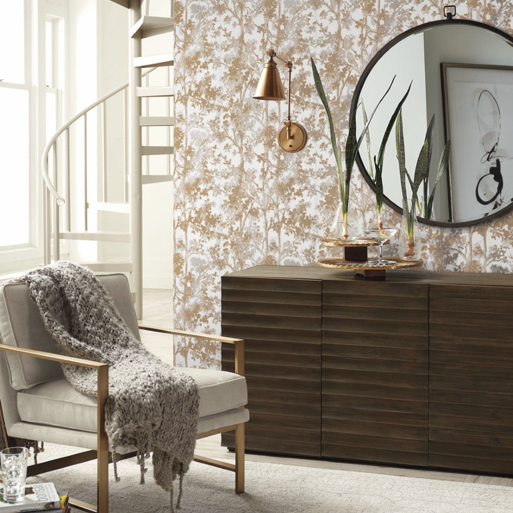 A cozy living room corner features a modern armchair with a gray knit throw. A dark wood sideboard is topped with a large mirror, plants, and decor. Elegance is enhanced by York Wallcoverings' White & Gold Shimmering Foliage Wallpaper and a spiral staircase.